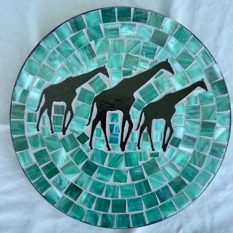 Round mosaic wall plate with three black giraffe silhouettes on a teal glass background