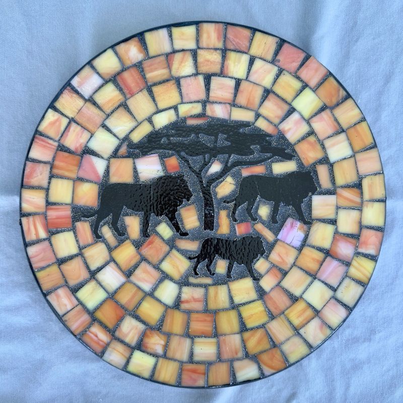 Round mosaic wall plate with three lion silhouettes beneath an acacia tree on a warm sunset mosaic of yellow, orange, and peach
