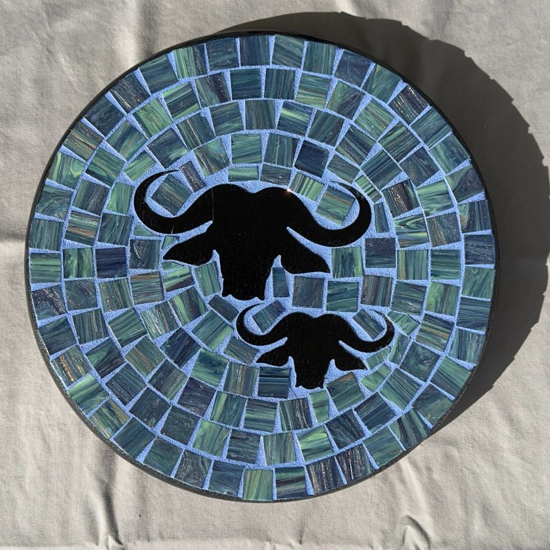 Round mosaic wall plate with two black Cape buffalo heads on a green and teal mosaic field with a lavender-blue inner ring