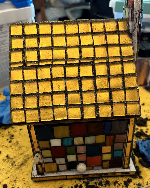 Small mosaic house with a gold glass roof and a multicolored checkered front, in progress on a workbench