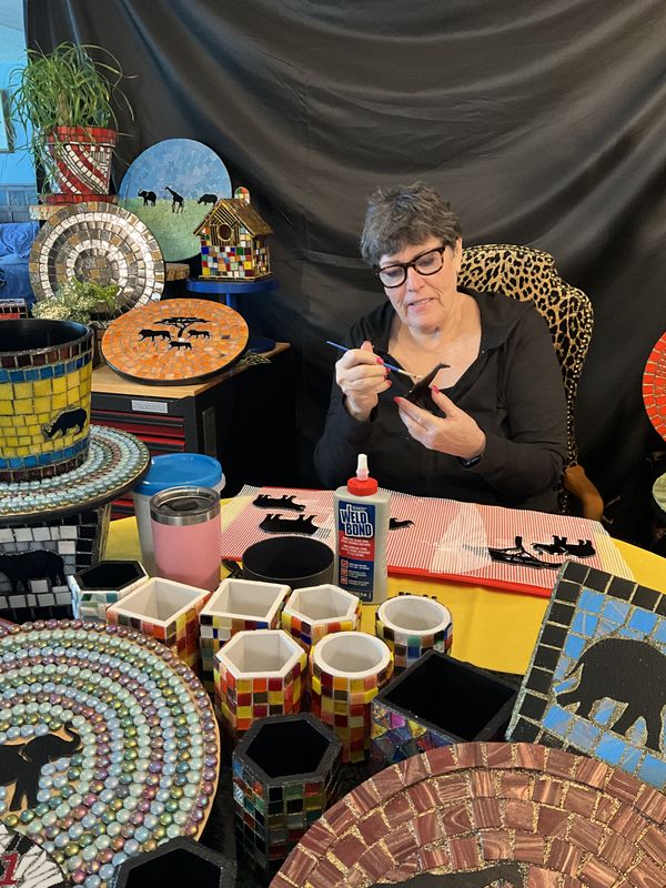 Janet at her studio worktable painting a small piece, surrounded by finished plates, planters, a birdhouse, and clocks