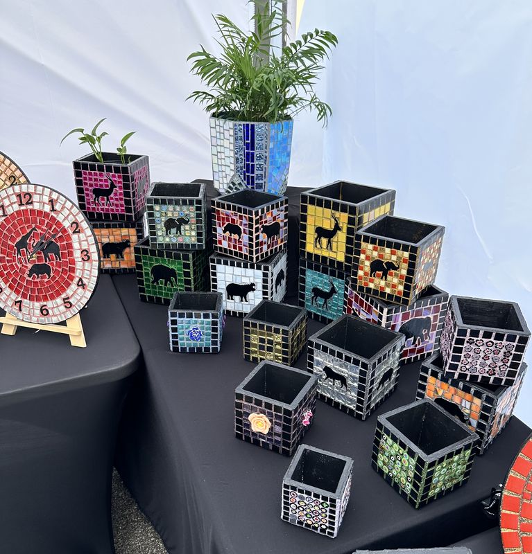 Craft-fair display with many square mosaic planters featuring animal silhouettes in assorted colors, plus a red mosaic clock on an easel