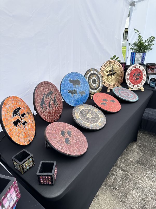 Craft-fair display with a row of round mosaic plates and clocks on easels and a foreground of small planters