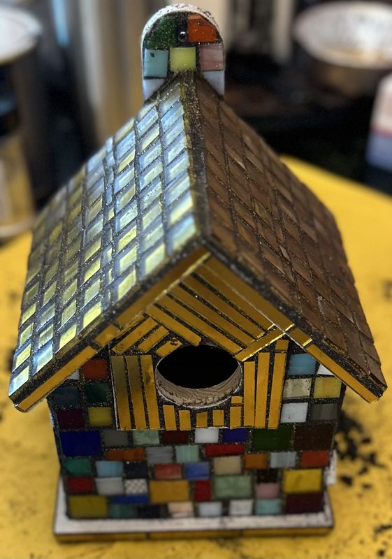 Small mosaic birdhouse with iridescent blue roof on one side and amber-gold roof on the other, with multicolored base tiles